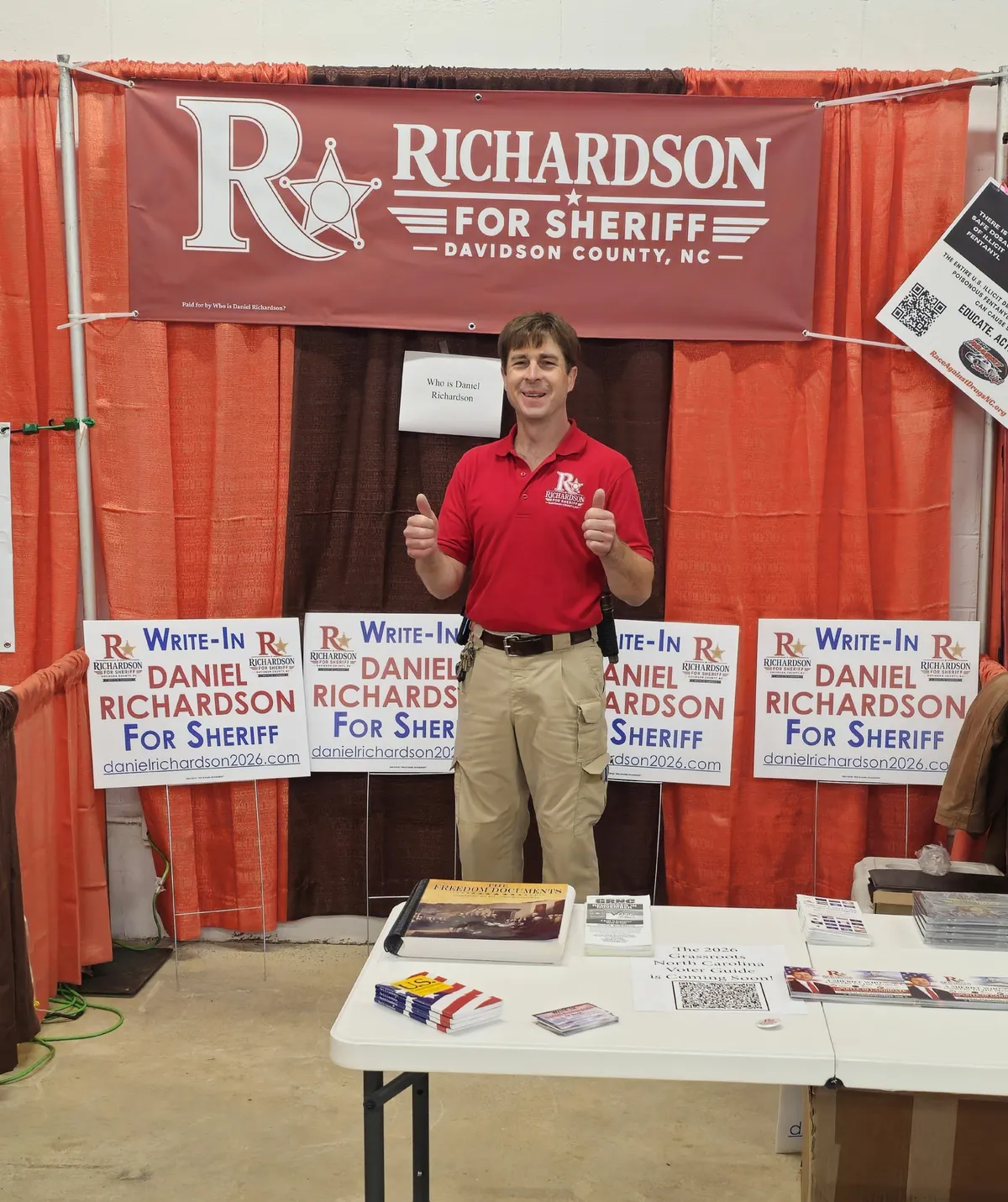 Daniel Richardson giving a thumbs up at his campaign booth, with Write-In Daniel Richardson for Sheriff yard signs and a Richardson for Sheriff banner behind him