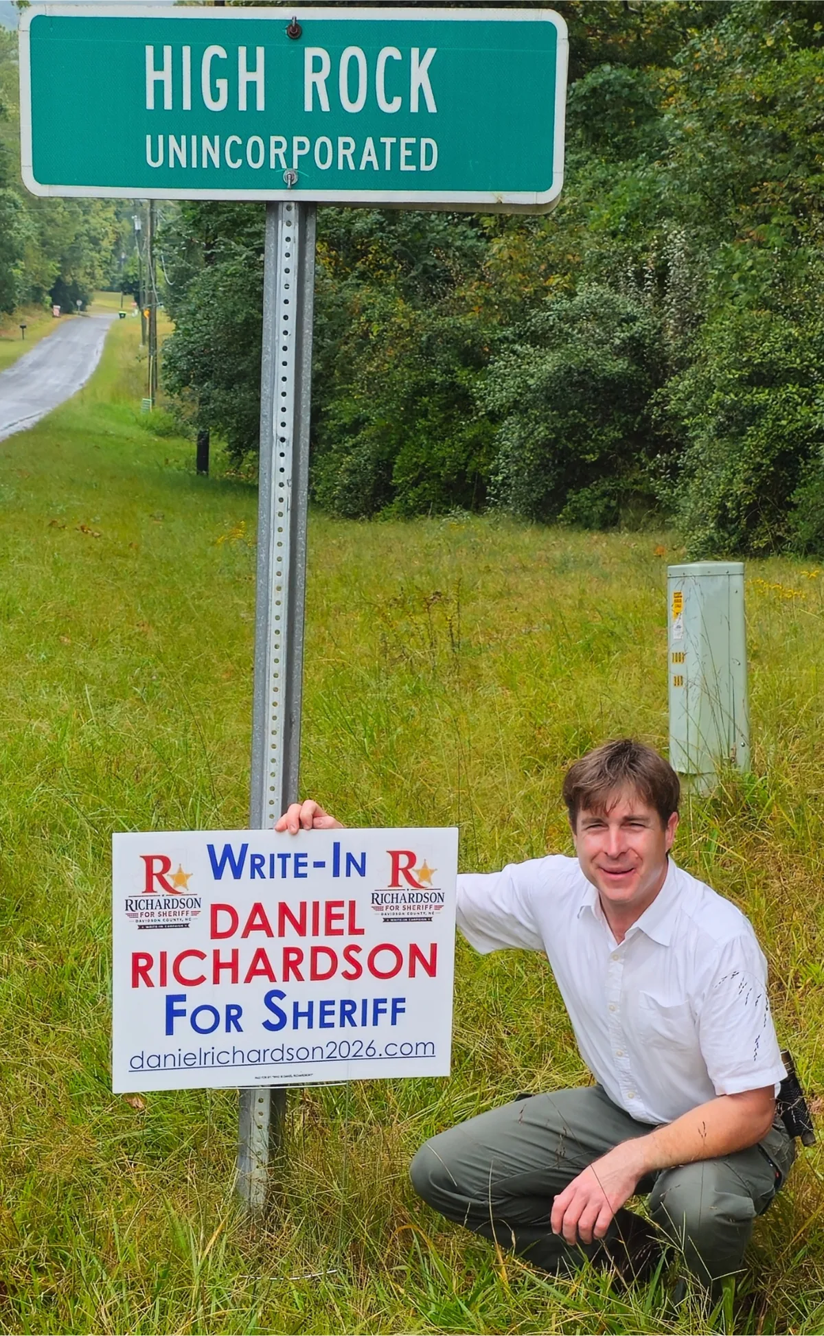Daniel Richardson kneeling beside a High Rock sign, holding a Write-In Daniel Richardson for Sheriff yard sign